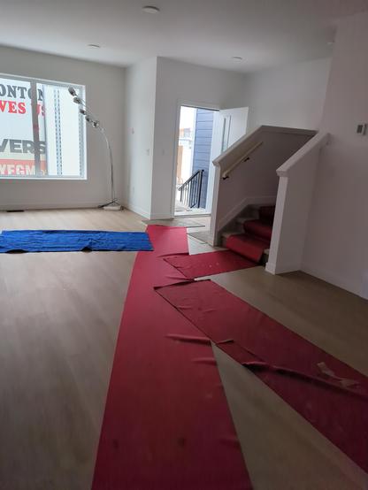 Empty, white walled open concept main floor with stairs up. Red carpet strips run through areas so that the movers can bring things in.
