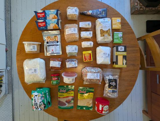 A top-down view of a collection of foods arranged neatly on a tabletop. Mostly dried ingredients like mashed potato flakes, coconut milk powder, but also spices and nuts and noodles and so on.