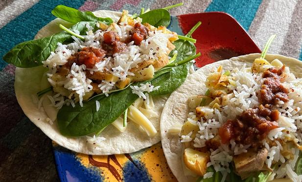 A colorful plate sitting on a table containing two tacos: flour tortillas, shredded cheese, green spinach leaves, diced roasted sweet potato, white rice, and topped with spicy tomato salsa.