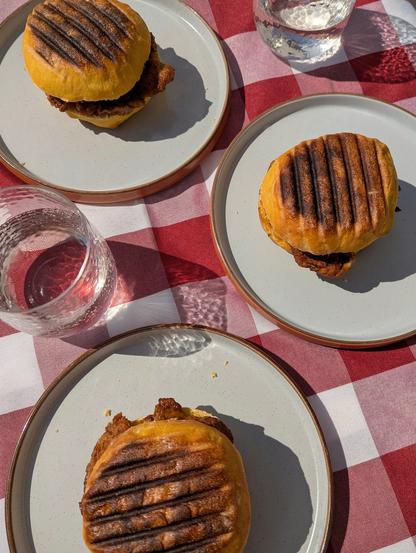 A table with food and drinks in the sunshine. There are three plates each with a bifana on them - an orange-coloured roll with brown griddle lines on top, containing slices of dark red-brown marinated pork. The drinks are all clear, and the glasses cast reflections on the table. There is a red and white check tablecloth over the table.