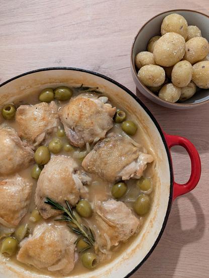 A photo of a pan containing a chicken thigh dish and bowl with potatoes, sitting on a wooden counter. The chicken thighs have golden skin tops, and sit in a creamy sauce with green olives and rosemary sprigs. The potatoes are small, slightly wrinkled, and have a very fine, intermittent white crust to them.