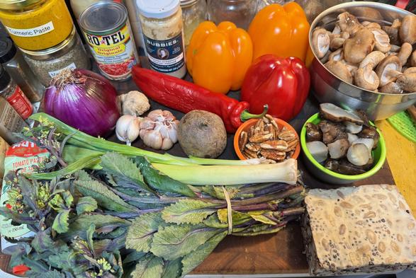 A cutting board on which are two jars (with homemade labels: turmeric paste and marjoram), a can Ro•Tel Original tomatoes, 1 jar Bubbies Horseradish, 2 orange bell peppers,1 red bell pepper, a steel bowl holding about a qart of small shiitake mushrooms, a large red onion, 1 bulb of red garlic plus another 1/4 bulb, 1 red swee-tooth pepper, 1 red beet, a small bowl of pecan halves, a bigger bowl of steamed pieces of Jerusalem artchoke, 1 small leek plus a scallion, 1 bunch red-kale sprouts, and a block of homemade tempeh.