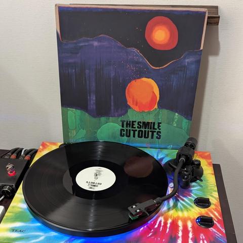 Un tocadiscos con un vinilo de The Smile Cutouts está sobre una plataforma con un diseño psicodélico de espiral multicolor. El vinilo es negro y se muestra la etiqueta blanca. La carátula del álbum muestra una escena nocturna abstracta con dos círculos anaranjados brillantes que parecen soles, sobre un paisaje oscuro y con pinceladas verdes en la parte inferior. El nombre de la banda está impreso en la portada.