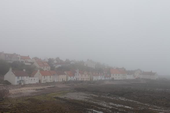 Colour photograph of a row of painted cottages fronting onto a sandy and rocky beach. Most of the picture is obscured by thick mist.