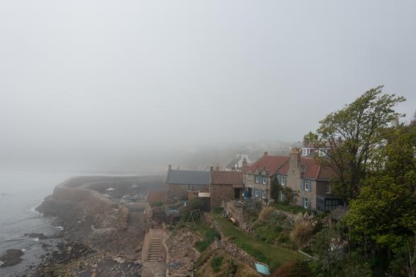 Colour photograph looking down on a small harbour surrounded by stone walls. There are several cottages fronting onto a tiny stretch of rocky foreshore. Everything beyond this is obscured by mist.