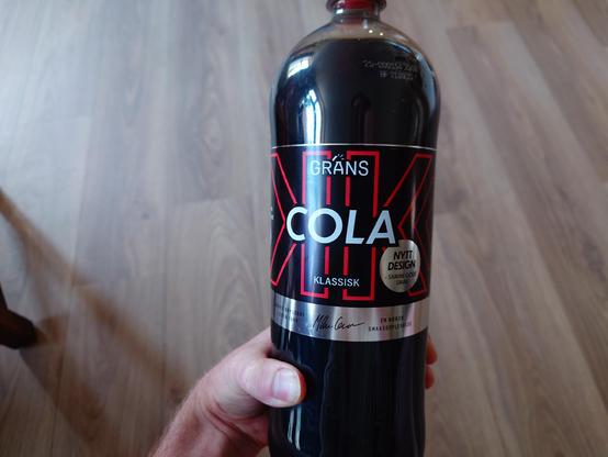 A photo of Matt holding the bottle of cola which now has an predominantly black label with red and silver highlights.