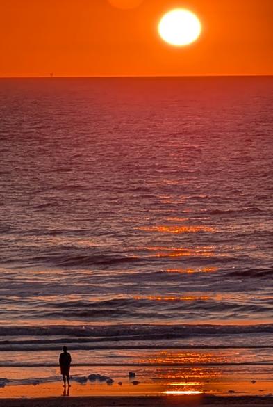 Fotofrafie, Sonnenuntergang am Meer, im Vordergrund die Silhouette eines Menschen, der in den Sonnenuntergang blickt. Orange Sonne, Spiegelungen auf dem Wasser, kleine Wellen