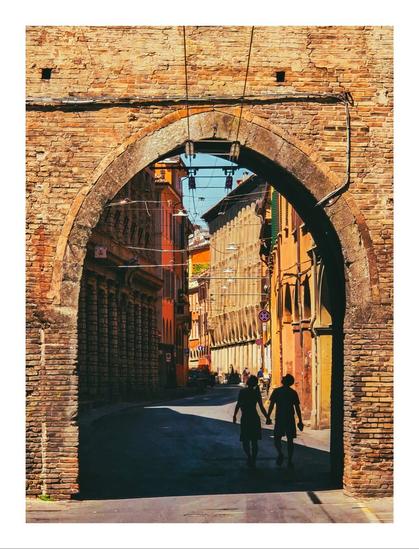 The photo is of an old  brick archway in via San Vitale in Bologna. Through the archway, there is a sunlit street lined with old buildings, ranging in colours from yellow to orange, to red. There are two silhouetted figures under the archway, that are walking hand-in-hand with their backs to us. They seem to be looking upwards at the building.