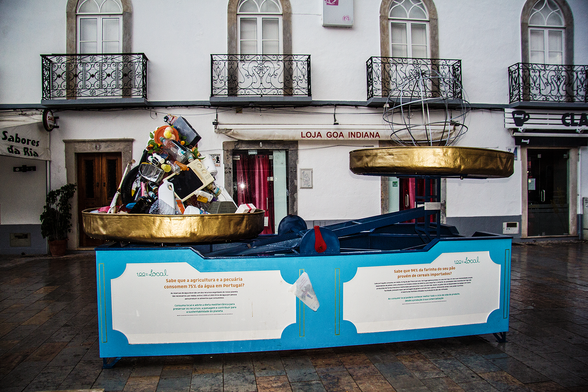 An installation in a pedestrian zone in Olhao in the Algarve, Portugal. It's a large weighing scale with all sorts of discarded items weighing down one side and a stylized terrestrial globe sculpture on the other. Panels on the installation's sides highlight issues around resource consumption and planetary sustainability; the two panels visible in the photo highlight water consumption for agriculture and food miles.