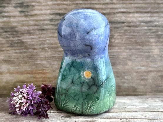 against a background of grey-brown driftwood, a raku ceramic earth spirit sculpture glazed in spring green, forest green, and blue (bottom to top). she has plant shapes and flowers carved into her and a yellow sun carving too.
