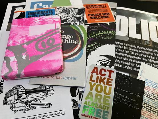The book and all of the stickers, zines, and posters laid out in a large pile. It’s colorful and there are lots of illustrations and different styles of typography. Illustrations include things like a Medusa head, an upside down tank, protest images, and skeletons. The snippets of visible text include “police not welcome,” “abolish class,” “anarchist appeal,” “act like you are already free,” and “immigrants welcome.”