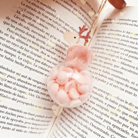 Sobre un libro abierto una bola decorativa de navidad con un reno rosa encima y melts pequeños rosas dentro. 