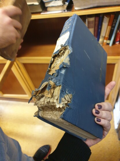 Libro de encuadernación en tela azul con el lomo reventado y la esquina inferior marcada por la trayectoria de una bala. 