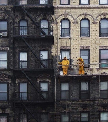 Dos operarios subidos en un andamio limpian la fachada de un edificio en NY. La parte sin limpiar es negra, la parte limpia de color geige claro.