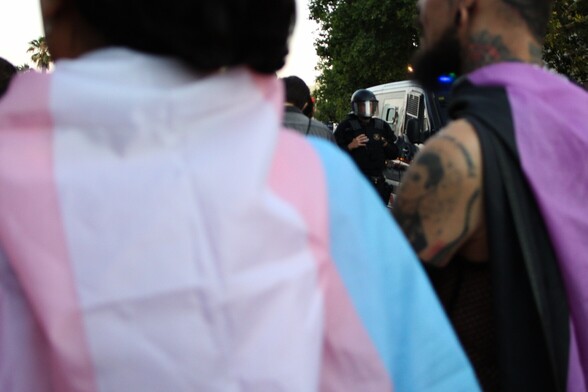 Y en primer plano hay una persona con una bandera trans sujeta en la espalda, y detrás por encima de su hombro se ve a un antidisturbios y una furgoneta de los Mossos d'Esquadra