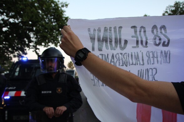 Primer plano de una mano sosteniendo una en primera fila de la concentración. En la esquina de la pancarta está escrito "28 de juny día per l'alliberament sexual y de genere". 
A la izquierda de la foto muy cerca de la pancarta hay un antidisturbios con el casco puesto y detrás un furgón de Mossos d’Esquadra 