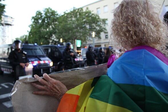 En primer plano vemos de espaldas a una mujer con la bandera arcoiris sujeta a la espalda, sosteniendo una pancarta frente a un montón de antidisturbios y furgonetas de los Mossos d’Esquadra 