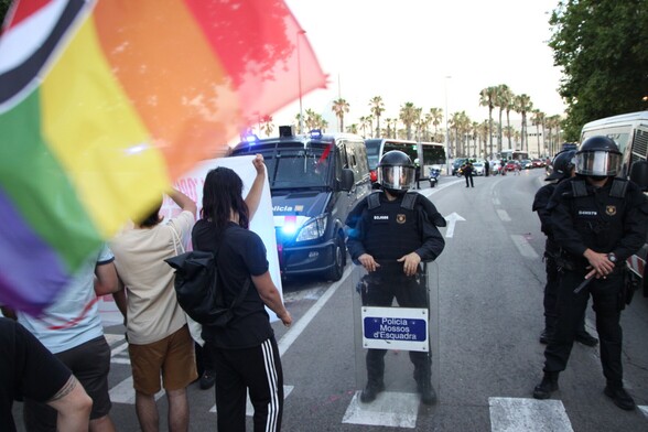 Varias personas sujetan una pancarta en primera fila de la concentración frente a los antidisturbios y las furgonetas de los Mossos d’Esquadra, de fondo se ve el atasco de coches provocado por la fila de Mossos que han impedido avanzar la manifestación antifascista.
En la esquina superior izquierda vemos un trocito de una bandera del orgullo de acción antifascista.