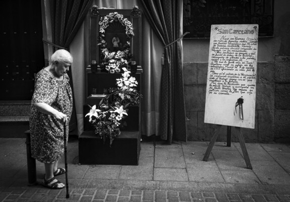 Foto en blanco y negro de una señora mayor paseando apoyada en un bastón por delante del altar de San Cayetano (junto a un manusescrito explicando el origen del mismo) en la Calle del Oso, Madrid