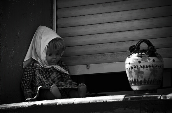 Foto detalle en blanco y negro de una muñeca chulapa junto a un botijo decorando una ventana (con la persiana bajada) por las fiestas de San Cayetano en la Calle del Oso, Madrid.