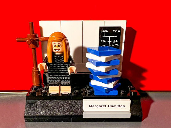 A Lego version of the famous Margaret Hamilton photo, next to the stacks of code she wrote for the Apollo missions. It includes the back wall, the blackboard with code and the coat rack.