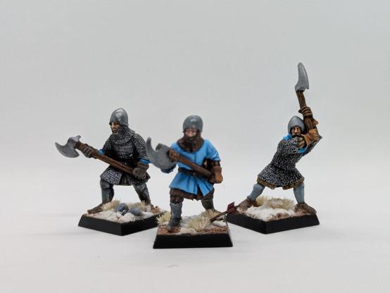 Tres hombres de armas con armadura de cota de malla dos de ellos y un uniforme azul el otro. Los tres blanden hachas a dos manos y caminan sobre nieve