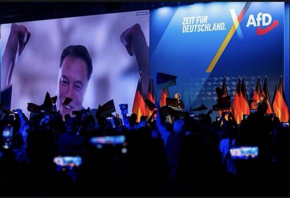 En una pantalla gigante sobre un escenario se proyecta la cara de Elon Musk levantando los brazos. En una maravillosa coincidencia, una de las banderas que ondea el público se coloca bajo la nariz de Musk, haciendo que parezca un bigote hitleriano.