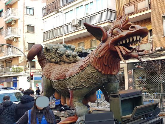 Una escultura de dragón de madera marrón y verde. Tiene una cara fiera de la que parecen salir lengyas de fuego, y cola doble. Llama la atención que lleva mamas protuberantes, como las de la tarasca española.