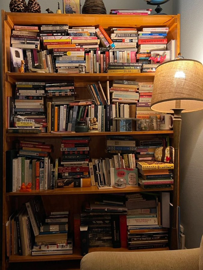 Estantería de madera clara petadísima de libros sin orden ni concierto casi metidos a presión con un montón de adornitos dispares como piñas, tazas, , fotos, figuritas y una bola del mundo en tó lo alto junto a una lámpara de pie torcida que arroja luz cálida a la escena. 