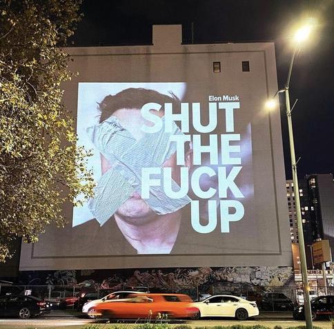 In a street, a large advertising board with a face of somebody that could be Elon musk, masked by adhesive tape. And on the right, written in block letters, 
Elon Musk
SHUT
THE 
FUCK
UP