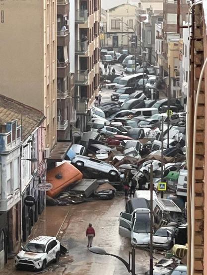 Photograph taken in Alfafar, Valencia, Spain, showing a city street littered with overturned and piled-up cars after massive floods caused by heavy rainfall. The road is covered in mud and debris, and a few people walk through the wreckage amidst buildings that appear largely intact. The scene highlights the extensive destruction and disruption caused by the flooding.