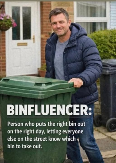 A white male dressed in dark winter coat, t-shirt  and jeans, smiling,  while he is  steering a large green garbage bin past a black one, as he walks it to the curb.
Caption : Binfluencer: Person who puts the right bin out on the right day letting everyone know which bin to take out.