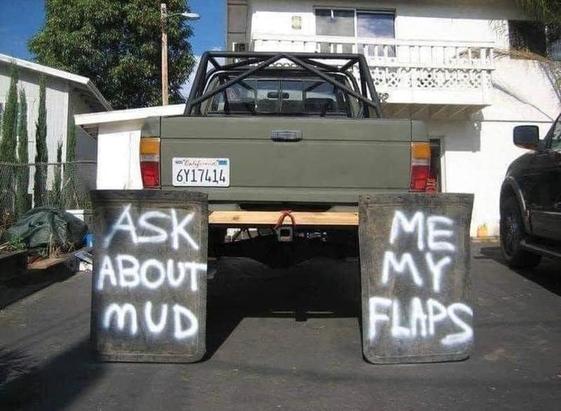 A very lifted truck with enormous mud flaps, probably repurposed from highway construction equipment. They are attached to a 2x4 that is in turn bungee corded to the trailer hitch. They've been spraypainted, one with "ASK ABOUT MUD" and the other with "ME MY FLAPS"