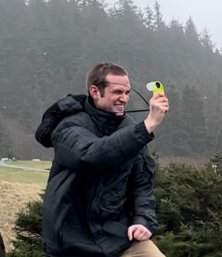 A photo of me, a late-20s white man, with a big grin while I struggle to stand in 80-100 km/h winds holding a handheld anemometer. The backdrop is a park surrounded by forest.
