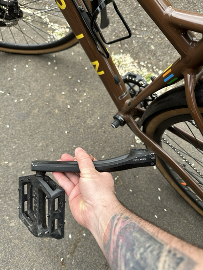 A bicycle missing its left crank and pedal in the background. A hand is holding the crank and pedal in the foreground. You can barely see that the area where the crank should attach to the crankset is broken.