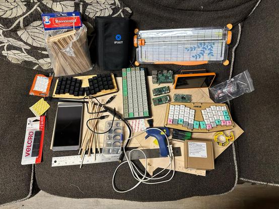 A pile of assorted computer parts, circuit boards, batteries, Keyboards, a ruler, paper cutter, and similar tools sitting on top of some pieces of wood on top of the couch.