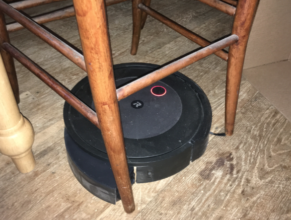 A robot vacuum cleaner under a chair. It's dust bin has opened, because the dust bin eject button is apparently something that can be pressed accidentally while the robot is moving around. It's a wonder why roomba just filed for bankruptcy.
