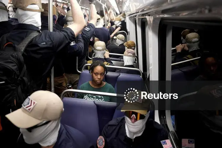 A lone Black woman sitting in a train car, surrounded by Patriot Front fascists too cowardly to show their face. Her expression is as you would expect in such a situation.