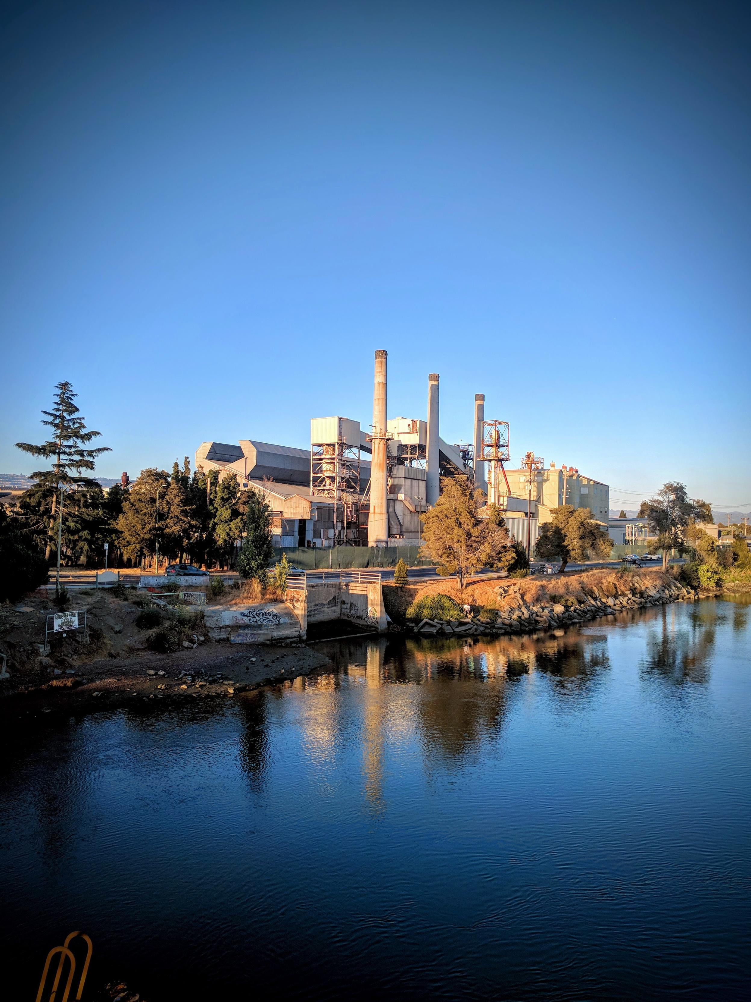 An old glass factory with a row of three smokestacks and various other industrial structures that are now rusted and silent. The photo is take from the fruivale bridge over the estuary and the factory is reflected in the water.