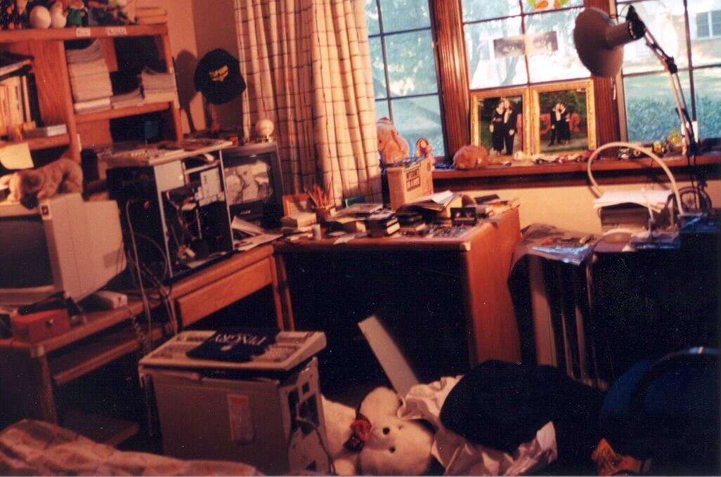 An L-shaped computer desk in my bedroom when I was ~19 with… way too much stuff. There’s two computer towers, one with an open side; a monitor and a TV. Nearly every inch is covered. There’s piles of stuff on the floor, bookshelves. A table is visible on the right edge with yet more stuff. 