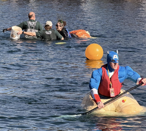 A man dressed as Captain America paddling in a giant pumpkin floating in a river, away from another pumpkin boat which has capsized where a paddler wearing an antler crown is being assisted by two volunteers