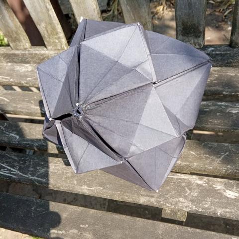 An origami polyhedron folded in grey-blue paper sitting on an old wooden bench. The model takes up most of the image. The shape is basically a cube with a pyramid on each face.