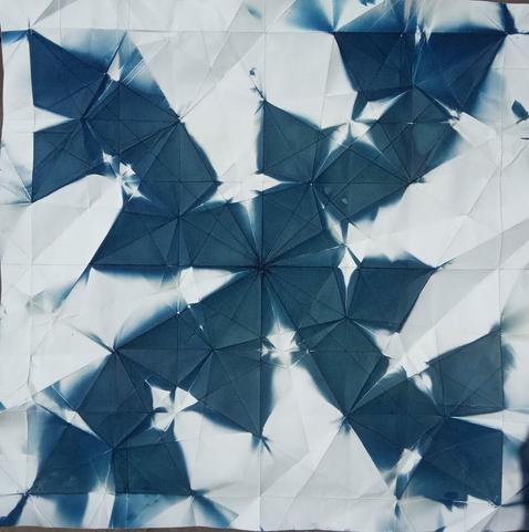 Square image, a geometric, symmetrical pattern of blue diamonds on a white background. Four tendrils of diamonds swirling away from the centre. The background is white.