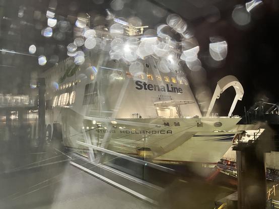 The bow of the Stena Hollandica from the passenger walkway on a draft rainy evening. Lights reflect off raindrops on the glass, causing a bokeh effect across the top of the ship. The bow is open to load vehicles.