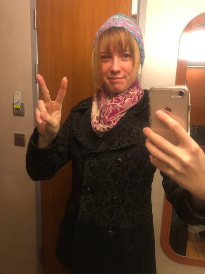 Mirror selfie in my hut! I'm wearing a black coat, a pink scarf with flowers and a blue-and-pink crochet hat. I'm making the V-shaped peace sign with my hand and looking in the camera with a slightly skeptical and massively tired look.