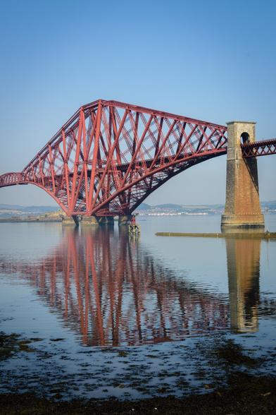 EN: View of the Forth Bridge from the waterside of River Forth. The railway bridge built of red steel is reflected on the water. The picture doesn't show the whole bridge but just the first third of it. 

DE: Blick von einer Terrasse am Fluss Forth hinüber auf die Forth Bridge. Die Eisenbrücke ist aus rotem Stahl erbaut und spiegelt sich im ruhigen Wasser. Das Bild zeigt allerdings nicht die gesamte Brücke, sondern nur das südliche Drittel.
