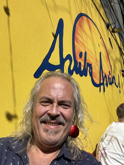 White haired man sits outside at a restaurant, showing over his shoulder the exterior wall with the restaurant name painted on it. Arriba Arriba.