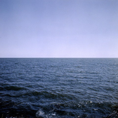 Blue of water is mirrored by blue of sky. Faint clouds and, barely perceptible, two sailboats ride the horizon line.