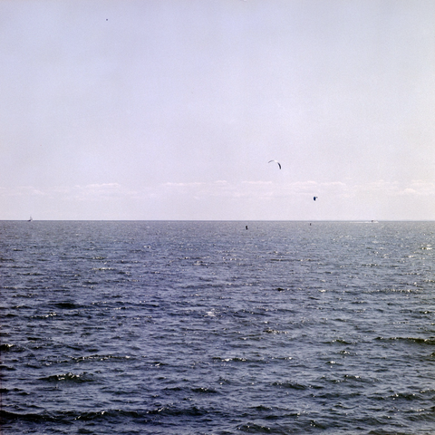 A deep blue water stretches to a pale blue sky, two parasails are drawn by a distant motor launch while a sloop rides the horizon line. A solitary bird flies far overhead.