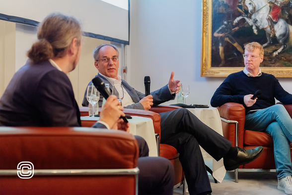 Das Bild zeigt eine Diskussionsrunde mit drei Personen, Prof. Dr. Christian Hummert, Forschungsdirektor der Cyberagentur, Uwe Cantner, Chef der EFI-Kommission und Marcvel Roth, Moderator und Journalist beim MDR. Sie sitzen in modernen Ledersesseln. Alle Teilnehmer halten Mikrofone, während einer aktiv spricht und die anderen zuhören. Im Hintergrund ist ein großes, gerahmtes Gemälde mit einer historischen Kampfszene zu sehen. Auf den Tischen stehen Wassergläser und Weingläser, was eine entspannte Atmosphäre andeutet. Das Logo der Cyberagentur ist unten links im Bild sichtbar. Foto: Nancy Glor/Cyberagentur